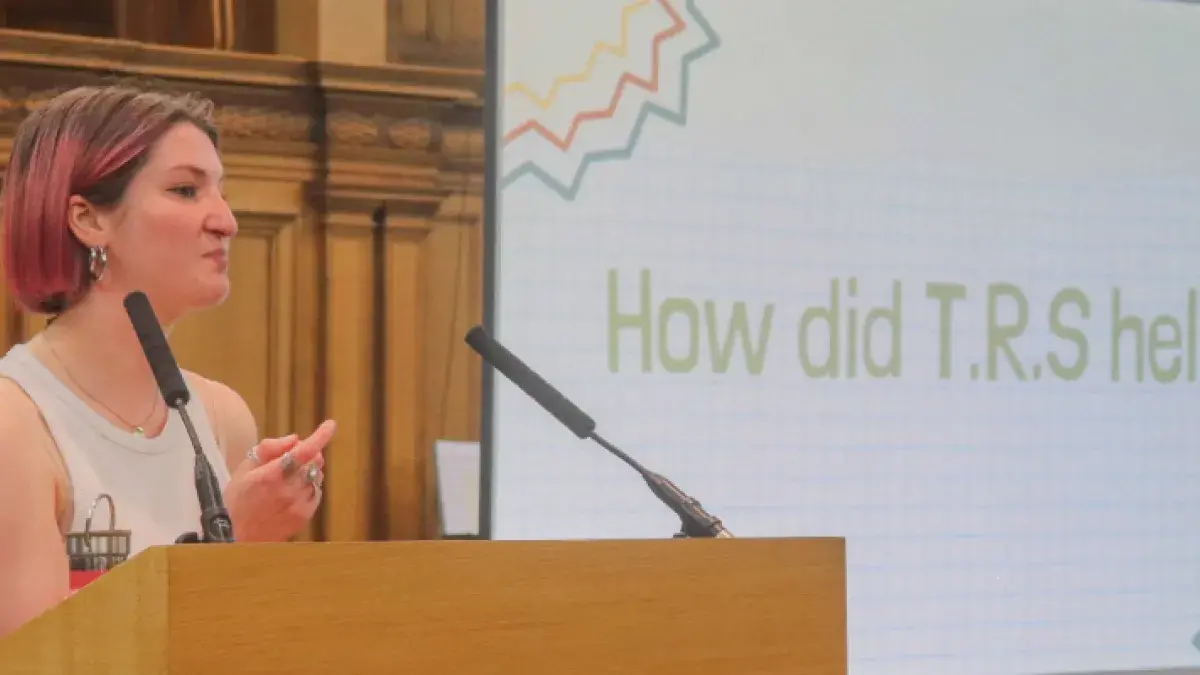 Colour head and shoulders photo of Beth Jones speaking at a lectern in the Assembly Hall. The words "How did T.R.S help?" are visible on a projector behind her.