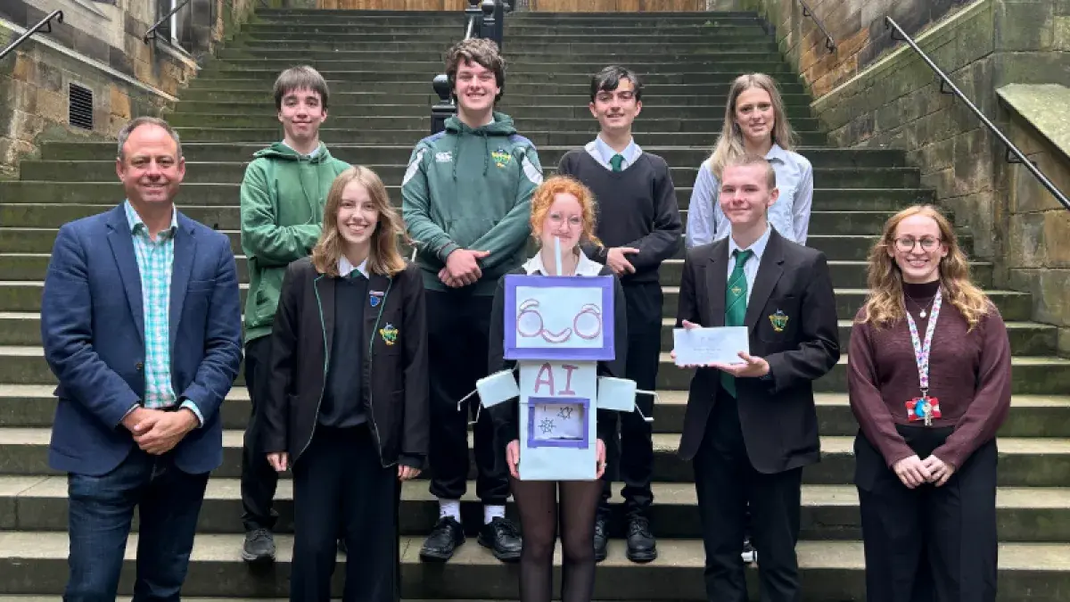 Colour head and shoulders photo of Dr Joshua Ralston stood smiling at the camera with the third place winners of the 2025 3 minute video competition as he hands them an envelope of their prize