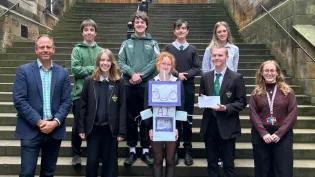 Colour head and shoulders photo of Dr Joshua Ralston stood smiling at the camera with the third place winners of the 2025 3 minute video competition as he hands them an envelope of their prize