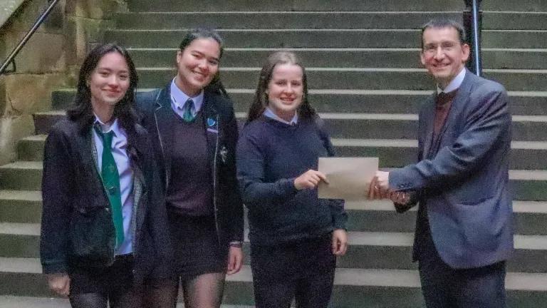 Colour photo of Professor Jeremy Carrette stood passing an envelope to competition winners Shona, Charlotte and Cecilia 