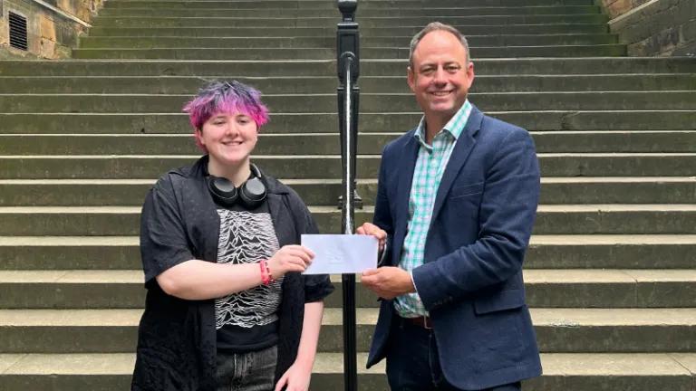 Colour head and shoulders photo of Dr Joshua Ralston stood smiling at the camera with the second place winner of the 2025 3 minute video competition as he hands them an envelope of their prize