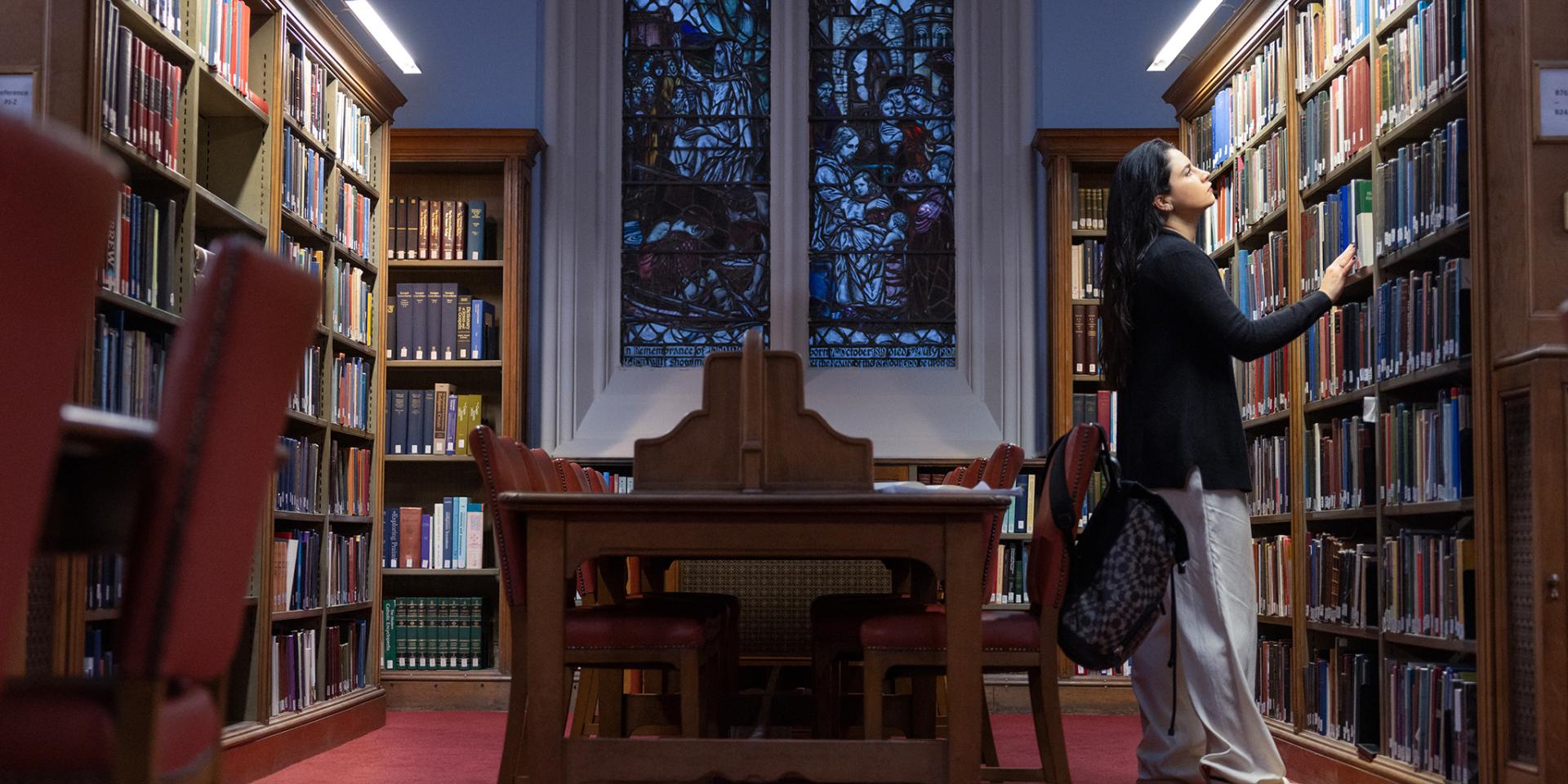 Colour photo of a School of Divinity student stood looking at books in the bookshelves in the New College Library