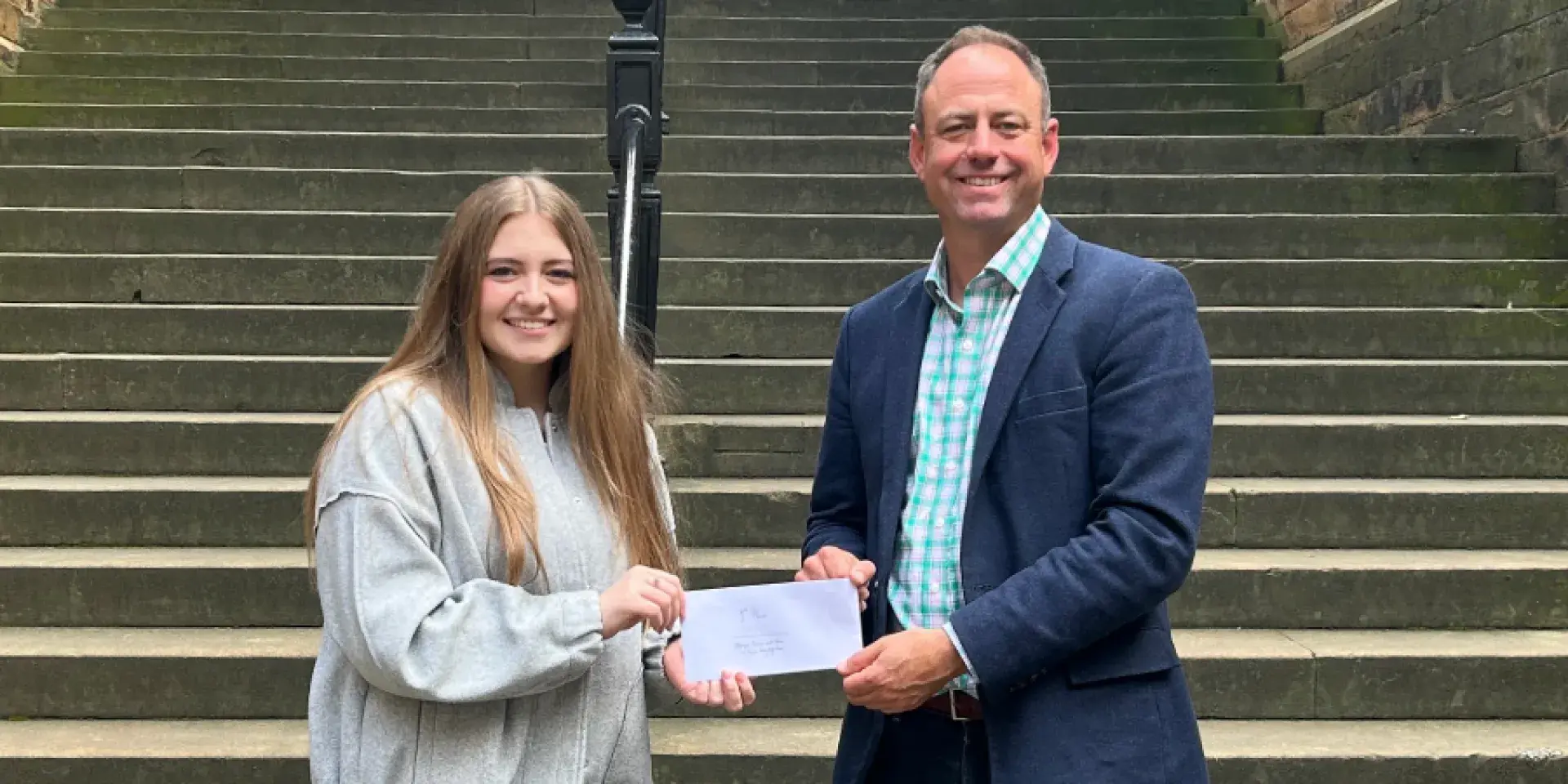 Colour head and shoulders photo of Dr Joshua Ralston stood smiling at the camera with the first place winner of the 2025 3 minute video competition as he hands them an envelope of their prize