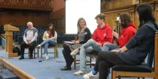 Colour photo of students and staff sat on the stage in the Assembly Hall as they talk to each other as part of a panel discussion