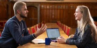 Colour photo of two students sat opposite one another in the Rainy Hall. They are smiling as they speak to one another.