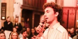 Colour head and shoulders photo of Sonnie Carlebach speaking into a microphone that they are holding