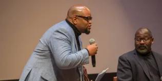 Colour head and shoulders photo of Eric Lewis stood speaking into a microphone that he is holding