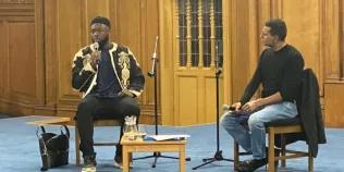 Colour photo of Imam Mustafa Briggs sat on a chair on the stage in the Assembly Hall whilst Dr Shadaab Rahemtulla sits beside him and listens