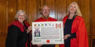 Colour photo of The Lord Lyon holding the New College Coat of Arms with Professor Susan Hardman Moore to his right and Professor Helen Bond to his left. They are all smiling.