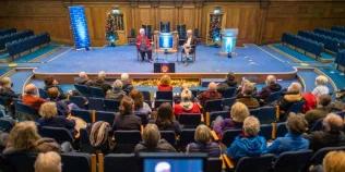 Colour photo of people sat in the Assembly Hall watching two people speak whilst sat on the stage