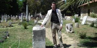 Colour photo of Sebastian Moll stood in a graveyard smiling at the camera as he rests his hand upon a stone with an inscription on it