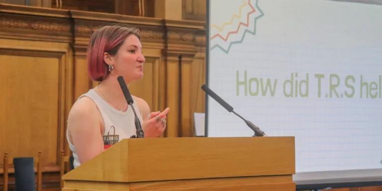 Colour head and shoulders photo of Beth Jones speaking at a lectern in the Assembly Hall. The words "How did T.R.S help?" are visible on a projector behind her.