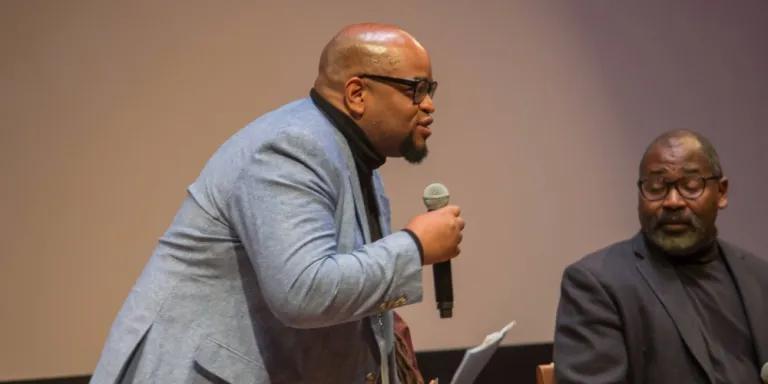 Colour head and shoulders photo of Eric Lewis stood speaking into a microphone that he is holding