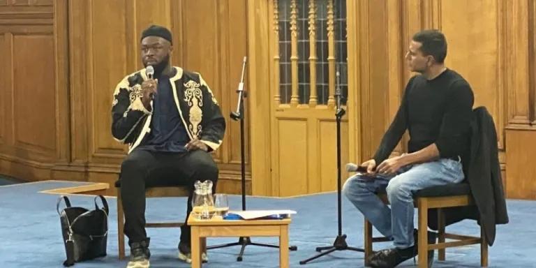 Colour photo of Imam Mustafa Briggs sat on a chair on the stage in the Assembly Hall whilst Dr Shadaab Rahemtulla sits beside him and listens