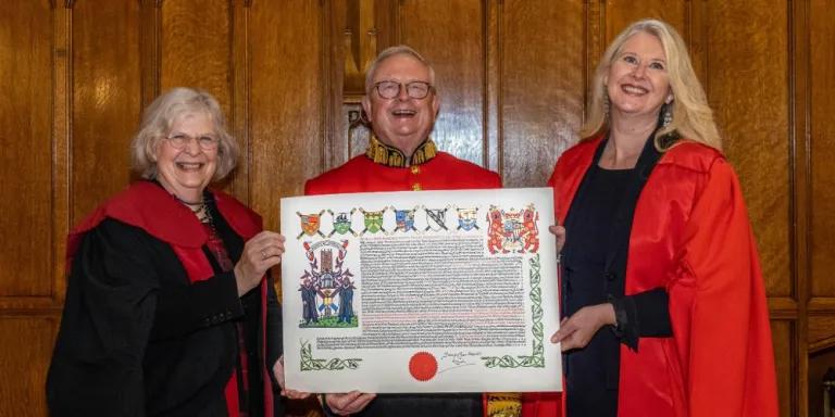 Colour photo of The Lord Lyon holding the New College Coat of Arms with Professor Susan Hardman Moore to his right and Professor Helen Bond to his left. They are all smiling.
