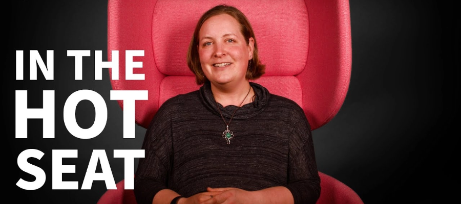 Colour head and shoulders image of Dr Bethany Sollereder sat in a red chair with a black background, smiling at the camera. White text to the side of Bethany reads, 'In the Hot Seat'