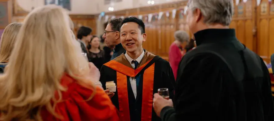 Colour photo of a School of Divinity Graduating Student stood talking to staff members in the Rainy Hall