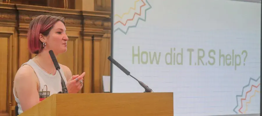 Colour head and shoulders photo of Beth Jones speaking at a lectern in the Assembly Hall. The words "How did T.R.S help?" are visible on a projector behind her.