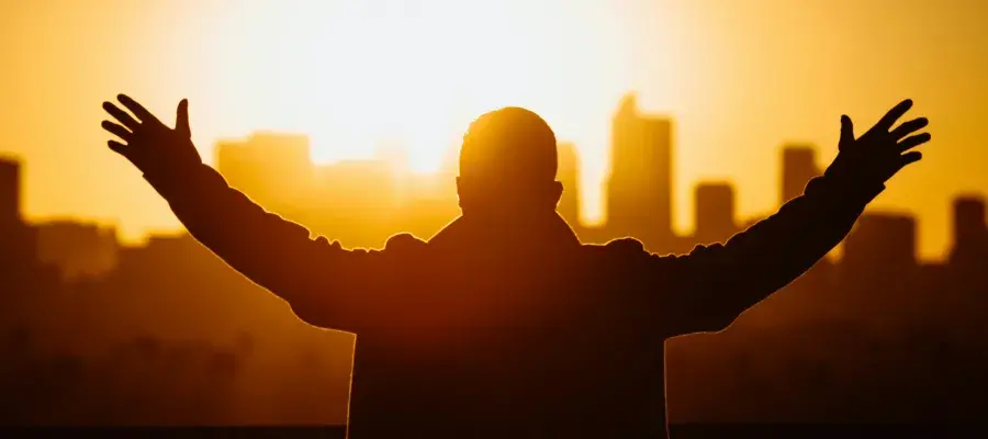 Colour photo of a man standing looking towards a city sunset with his arms outstretched and back towards the camera