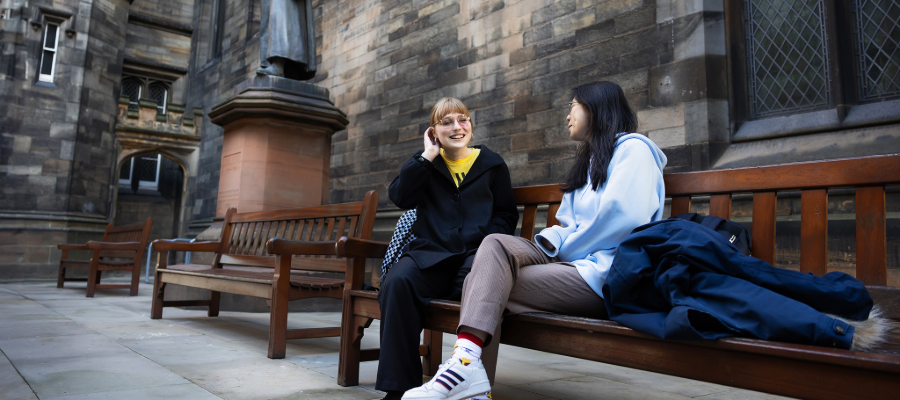 Colour photo of two students sat beside one another on a bench in the New College Quad. They are smiling and talking to one another.
