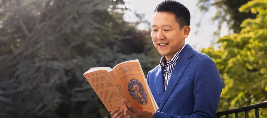 Colour photo of a student sat on a bench in a garden, reading a book