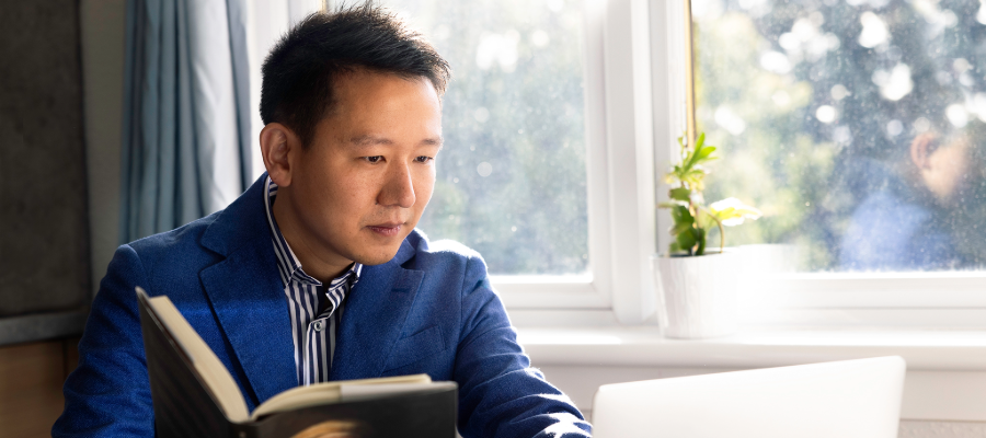 Colour photo of a student using their laptop at home whilst sat holding a book in their other hand