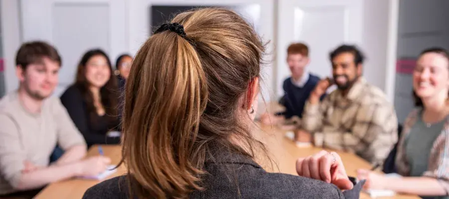 Colour photo of someone sat at the head of the table speaking to a group of students as they listen in. The photo is taken from behind the head of the speaker