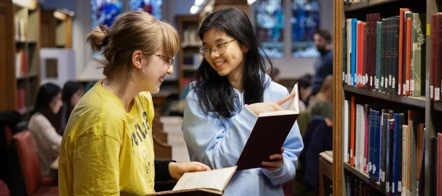 Colour head and shoulders photo of two students stood smiling together in the New College Library. They are both holding books