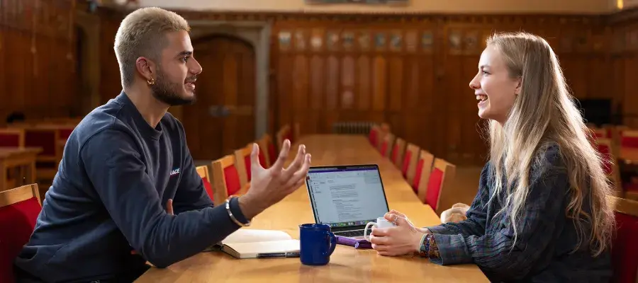 Colour photo of two students sat opposite one another in the Rainy Hall. They are smiling as they speak to one another.