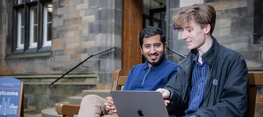 Colour photo of two students sat together in a bench in the New College Quad. The student sat close to the camera is showing the other student something on their laptop