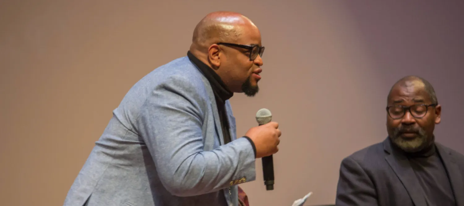 Colour head and shoulders photo of Eric Lewis stood speaking into a microphone that he is holding
