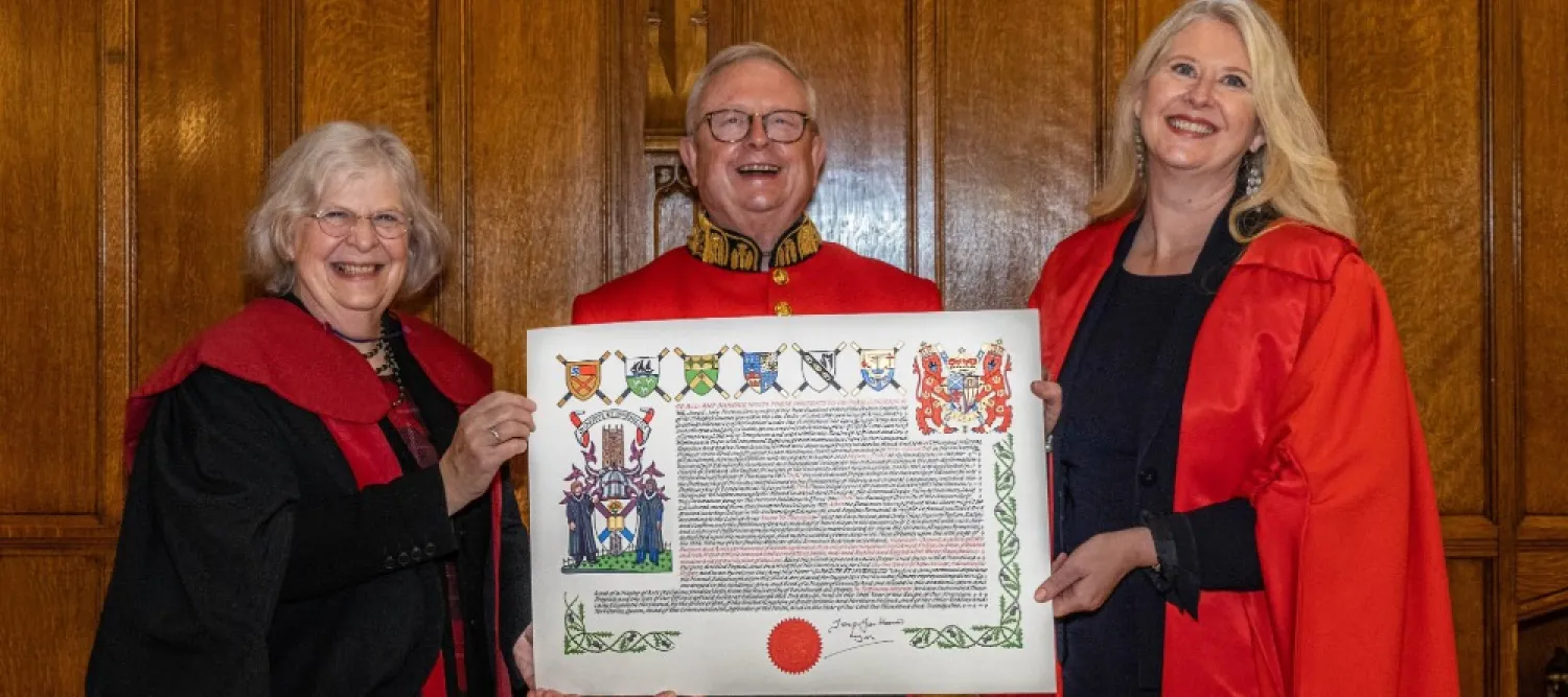 Colour photo of The Lord Lyon holding the New College Coat of Arms with Professor Susan Hardman Moore to his right and Professor Helen Bond to his left. They are all smiling.