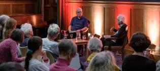 Colour photo of David Neville and Jenni Daiches sat together in the Martin Hall. David is looking to his left at Jenni as she speaks in front of the audience seated before her.