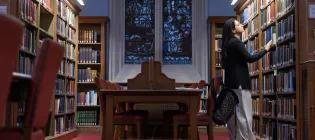 Colour photo of a School of Divinity student stood looking at books in the bookshelves in the New College Library