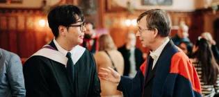 Colour photo of a School of Divinity Graduating Student stood talking to a staff member in the Rainy Hall