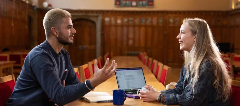Colour photo of two School of Divinity students sat talking at a table in Rainy Hall