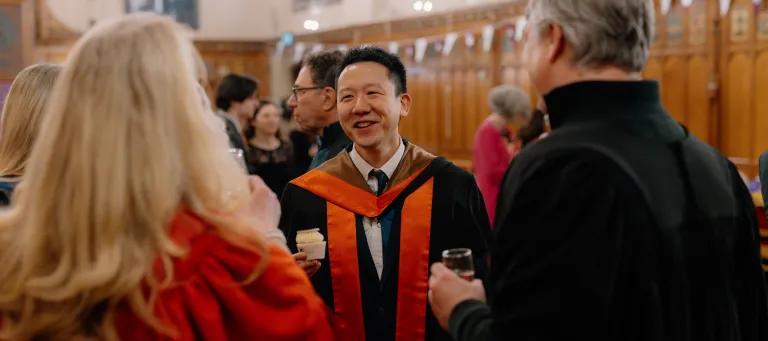 Colour photo of a School of Divinity Graduating Student stood talking to staff members in the Rainy Hall