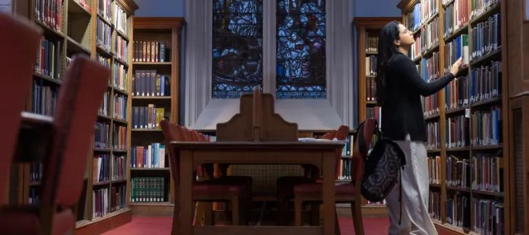 Colour photo of a student stood looking at books in the bookshelves of the New College Library