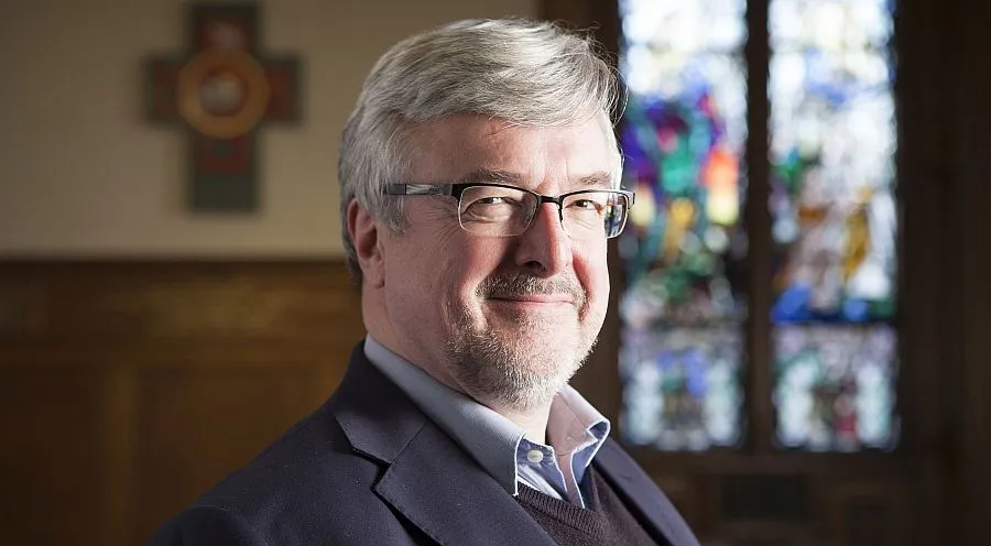 Dr Michael Fuller head and shoulders photo taken in the Martin Hall in front of a cross and stained glass window