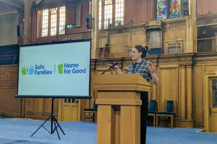 New College alumnus Tamsyn Radmall stands at a lecturn in Assembly Hall presenting a powerpoint presentation