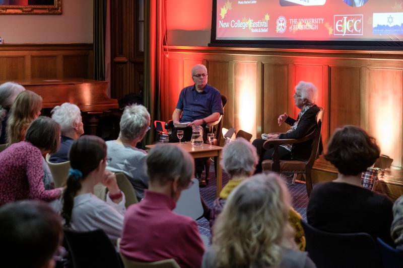 Colour photo of David Neville and Jenni Daiches sat together in the Martin Hall. David is looking to his left at Jenni as she speaks in front of the audience seated before her.