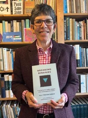 Colour head and shoulders photo of Elli Tikvah Sarah holding their book 'Beyond Binaries' as they smile at the camera