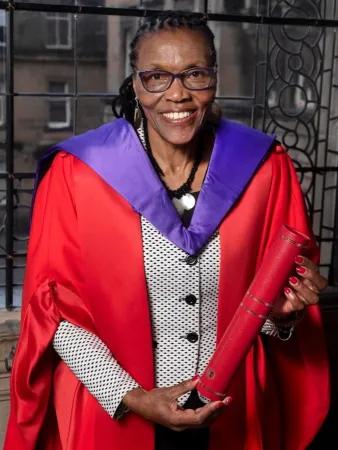 Colour head and shoulders photo of Esther Mombo smiling at the camera whilst holding her Honorary Doctorate